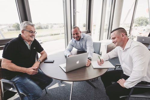 Drei Personen sitzen an einem runden Tisch und schauen auf den Bildschirm einen dem Betrachter mit dem Rücken zugedrehten MacBook